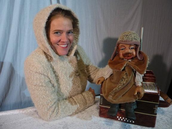 Schauspielerin Anke Scholz in einem hellen Wintermantel mit Kapuze hält Puppenfigur Wanja, der auf einem Akkordeon sitzt